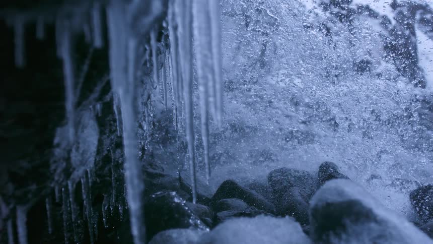 A frigid waterfall crashes over snow-covered rocks in stunning slow motion next to hanging icicles