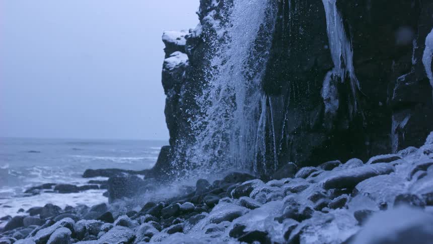 A waterfall cascades in slow motion over an icy cliff onto a snow-covered rocky beach next to the ocean