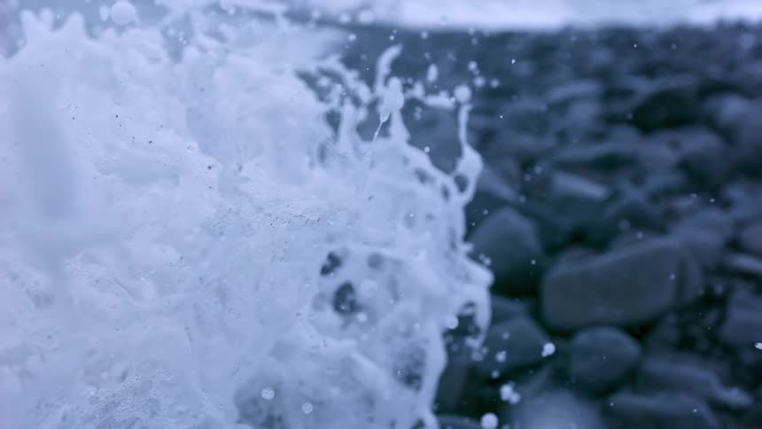 Foamy ocean wave splashes over dark rocks on the shore in extreme slow motion
