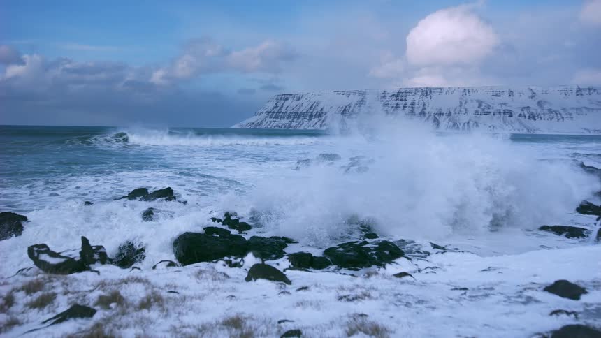 Powerful ocean waves crash in super slow motion against a snowy shoreline in front of a majestic winter mountain