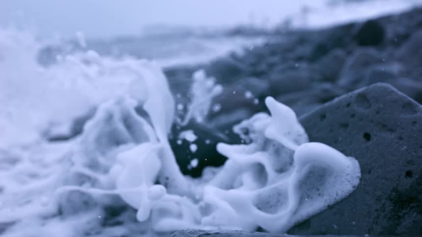 Foamy white ocean waves crash against dark rocks on the shore in dramatic super slow motion