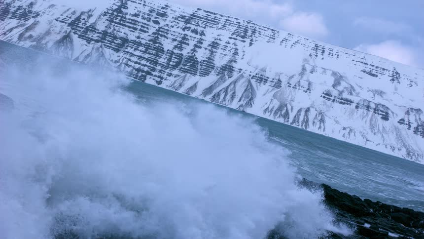Super slow motion of massive waves crashing onto a rocky shore with snowy Icelandic mountains in the background