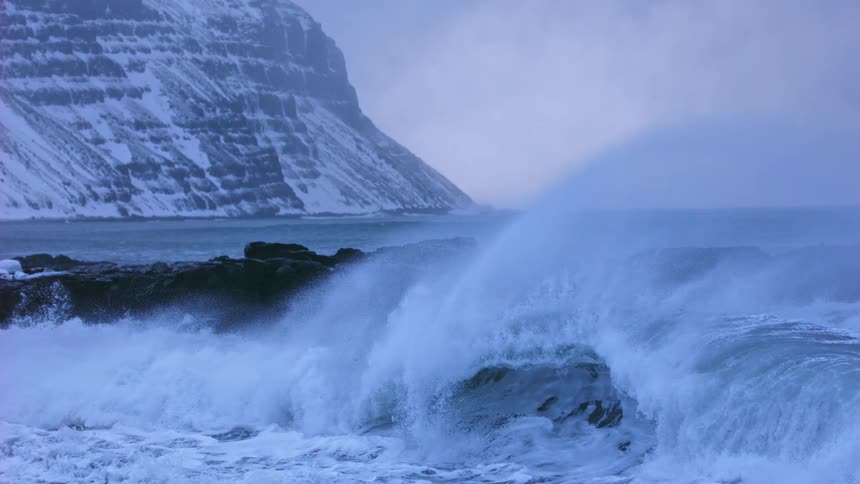Cinemagraph of a powerful ocean wave frozen in time as it crashes against rocks with a snowy mountain in the background