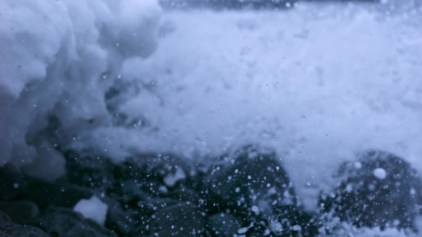 A wave crashes into snowy rocks on the shore in extreme slow motion