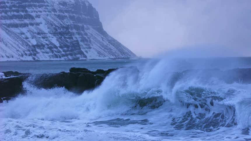 A dramatic cinemagraph shows a powerful ocean wave frozen in time as it crashes against a rocky, snow-covered coast