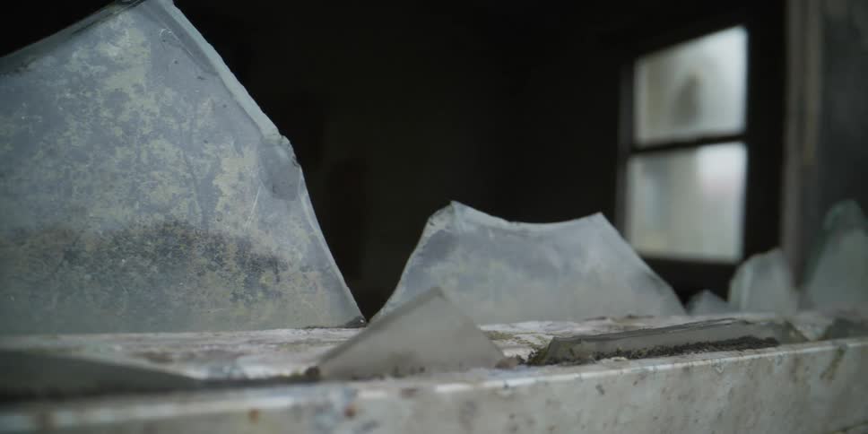 Close up slow pull back from sharp shards of glass in a decaying and broken window frame of an abandoned building