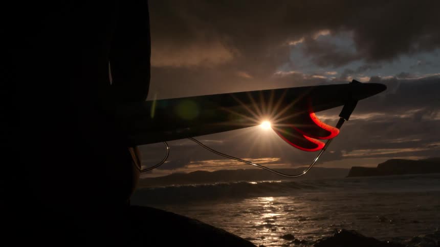A silhouetted surfer holds their board with a glowing red fin, watching the sunset over the ocean waves