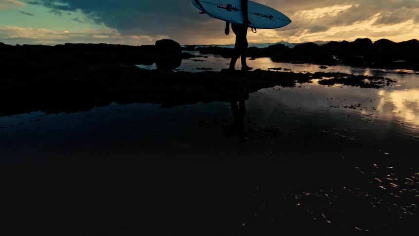 A silhouetted surfer walks with their board across a rocky shoreline at sunset, their form reflected in a calm tide pool