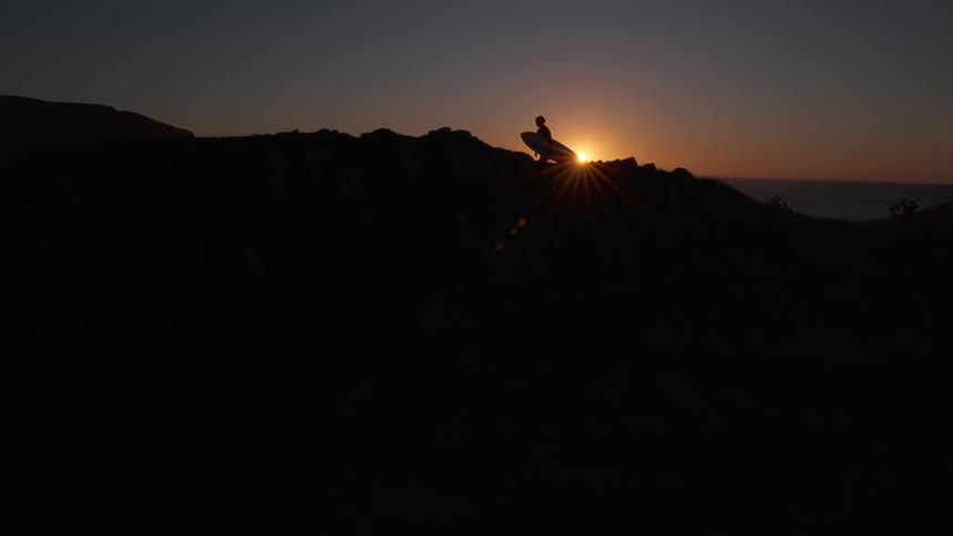A silhouetted surfer carrying a surfboard walks along a coastal ridge at sunset as the sun flare shines