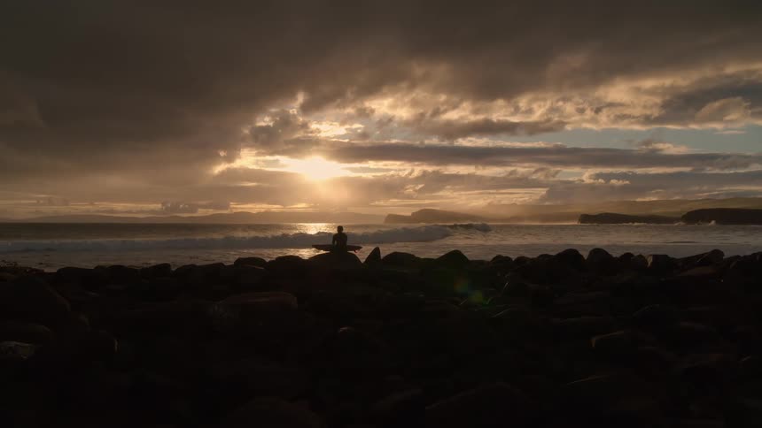 A lone surfer sits silhouetted on a rocky coastline, contemplating the ocean waves during a dramatic and beautiful sunset