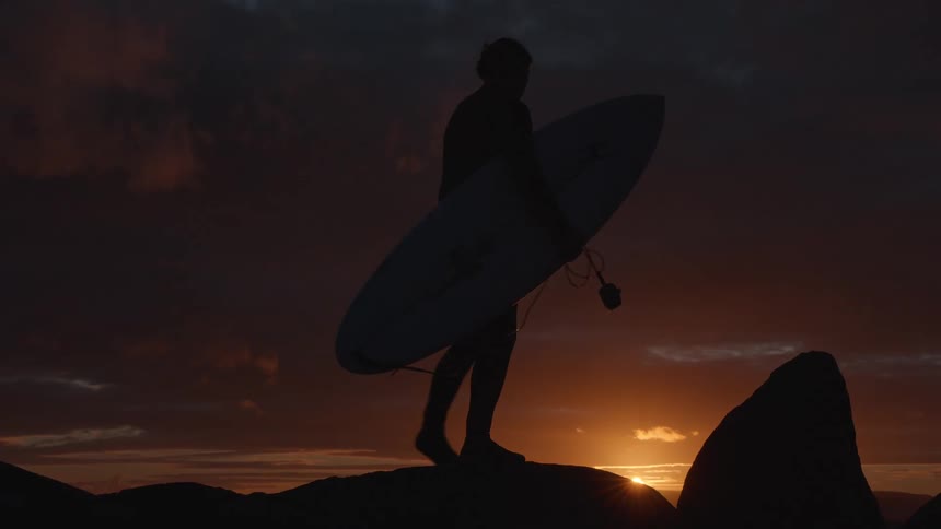 Slow motion silhouette of a surfer carrying a surfboard across rocks against a dramatic sunset sky