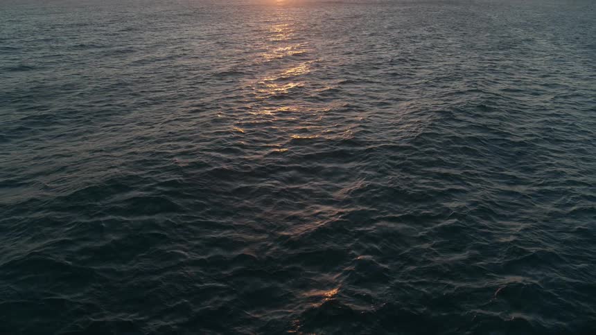 Flying backward over dark ocean waves reflecting the beautiful sunset on the horizon