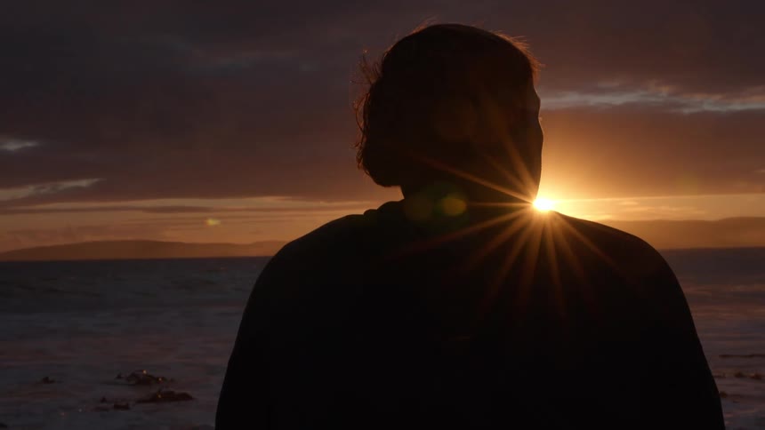 Static shot of a silhouetted person watching the sun crest the ocean horizon, creating a beautiful starburst effect