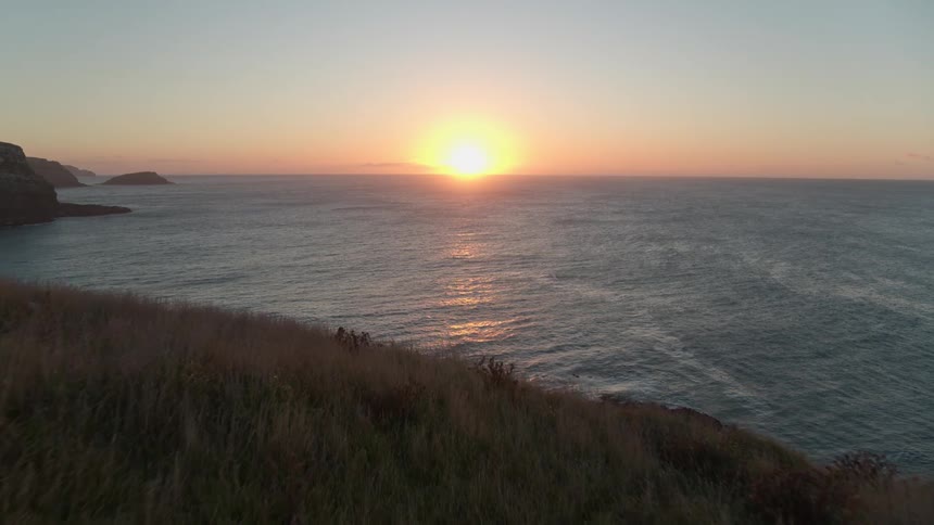 A beautiful aerial shot slowly moves over a grassy cliff to reveal the vast ocean and a stunning sunset on the horizon