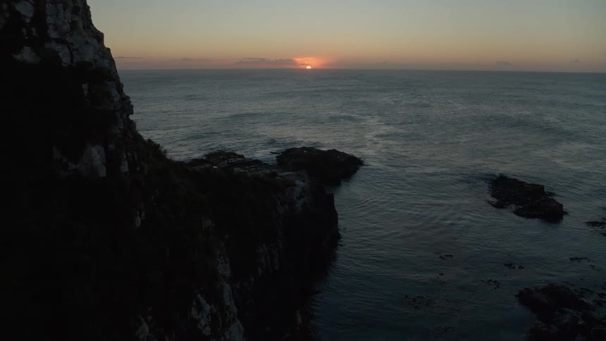An aerial view slowly glides over a dark, rocky coastline towards the ocean as the sun rises on the horizon