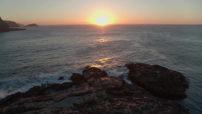 The sun sets over a tranquil ocean, its golden light reflecting on the water as waves crash against a rocky coastline