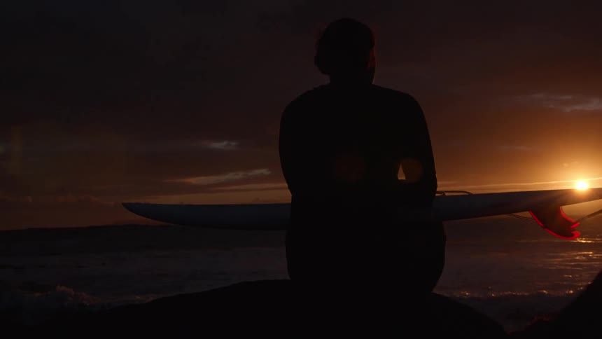 A silhouetted surfer sits with their board on a rock, peacefully watching the sun set over the ocean waves