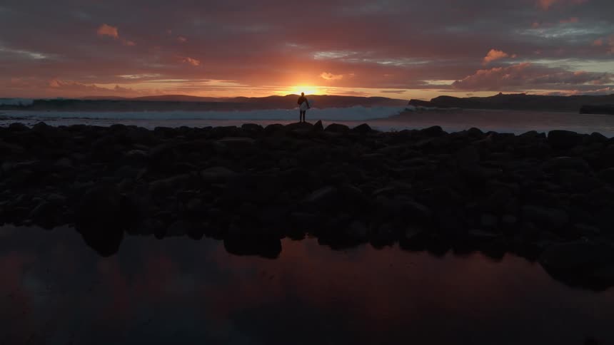 Dramatic aerial shot moving away from a lone surfer standing on a rocky beach, watching the waves during a beautiful sunset