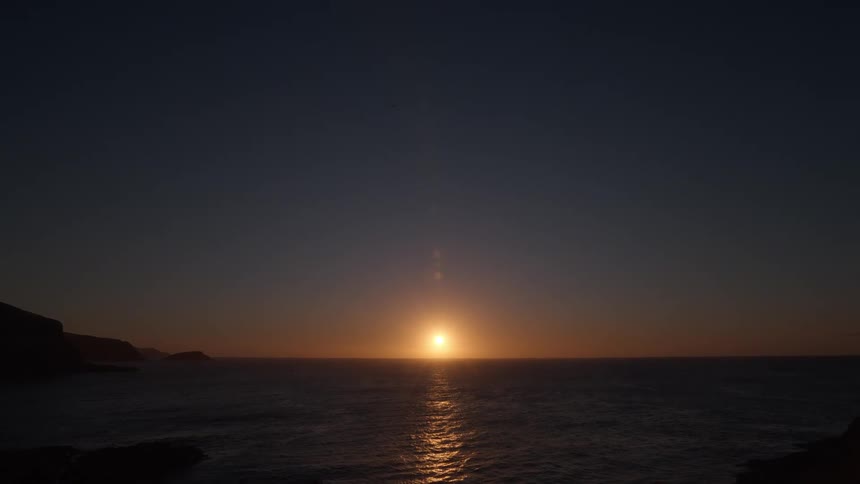 A beautiful golden sun sets over the ocean, viewed from a silhouetted rocky coast