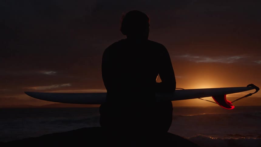 A silhouetted surfer sits with their board, peacefully watching the sun set over the ocean waves