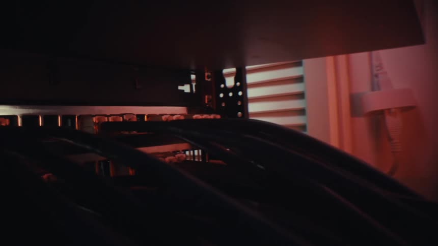 A close-up dolly shot pushes through a tangled mess of cables to a network switch in a dark server room lit by red lights