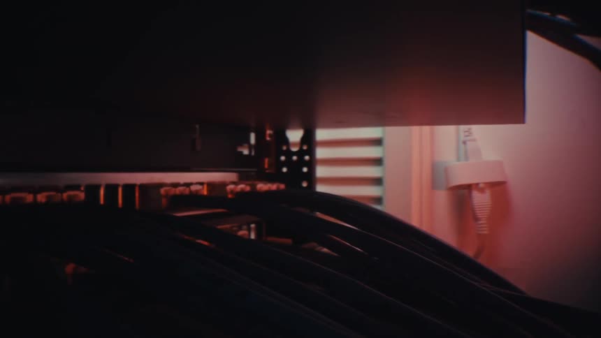 A moody dolly-in shot reveals the intricate network of cables plugged into a server rack in a dark data center