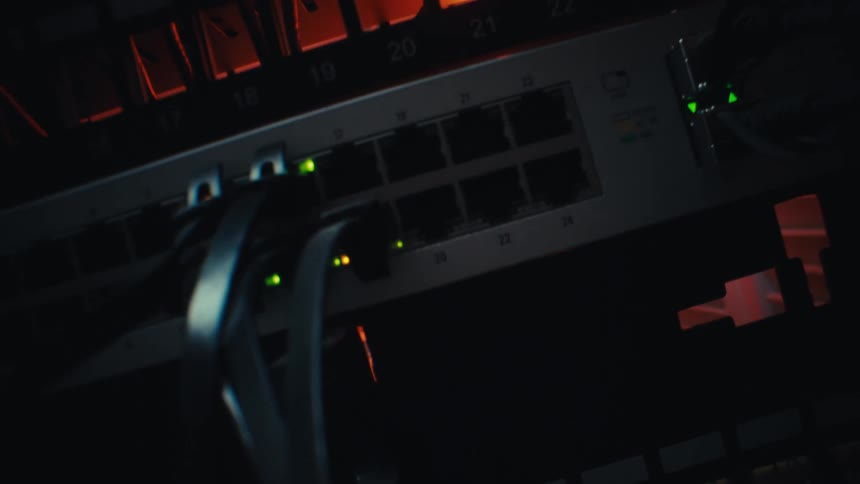 A dramatic close-up tilt shot reveals a working network switch in a server rack, with its lights blinking in a dark, red-lit data center