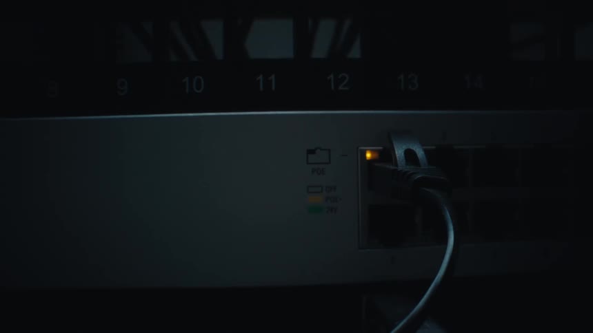 A close-up of a blinking yellow light on an ethernet port pulls back to reveal the network switch in a dark server room
