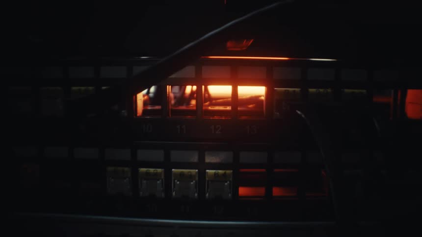 A cinematic slow pull-back shot reveals a network patch panel and cables backlit by a warm orange glow in a dark server room