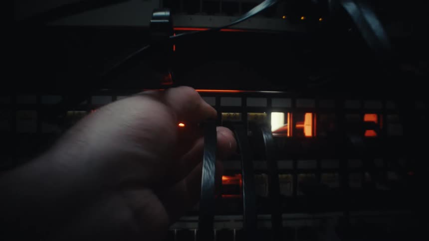 An IT technician's hand connects a network cable into a server rack patch panel in a dark, glowing server room