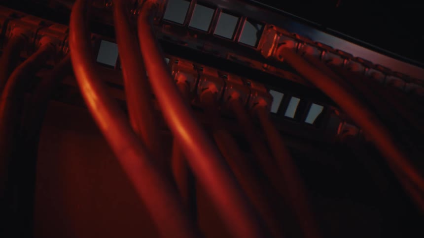 A close-up dolly shot of red internet cables plugged into a network switch in a dark server room under red alert lighting