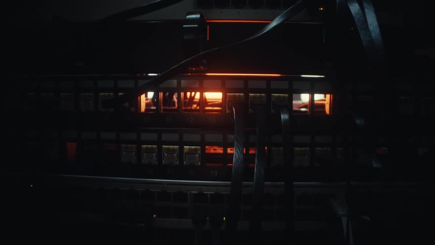 A network switch with connected cables glows with a warm, ominous light in a dark data center as the camera slowly pushes in