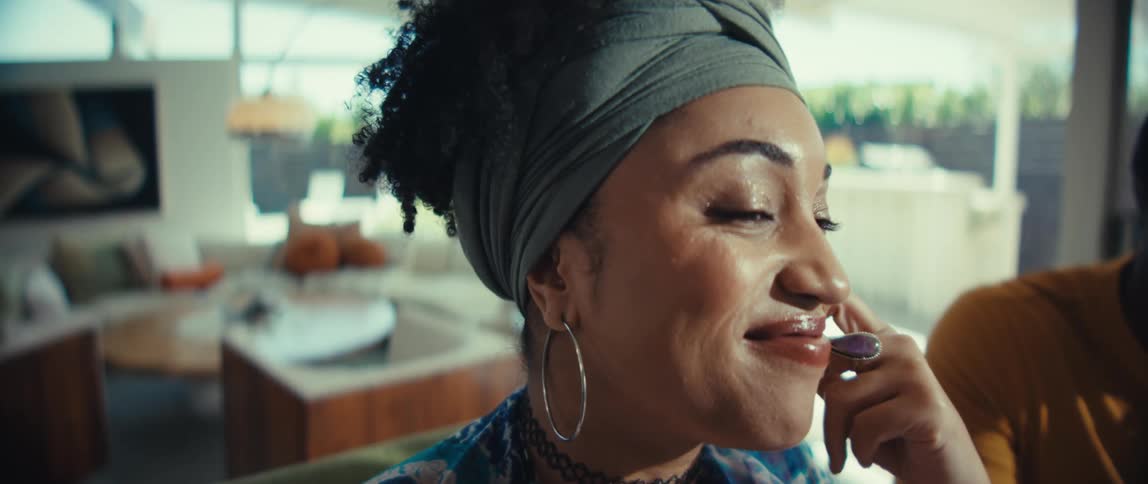 A close-up of a stylish woman with a headwrap smiling and listening thoughtfully in a sunlit modern home