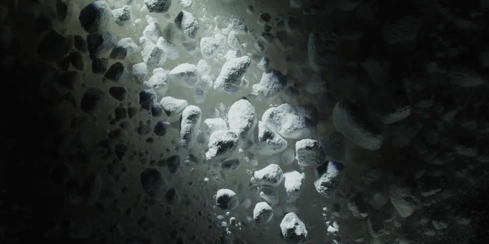 A beam of light sweeps across a dark surface, revealing a cluster of white rocky particles that erupt in a slow-motion, zero-gravity cloud
