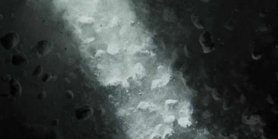 A dramatic beam of light illuminates fine white powder and rocks as they float and settle in slow motion against a dark background