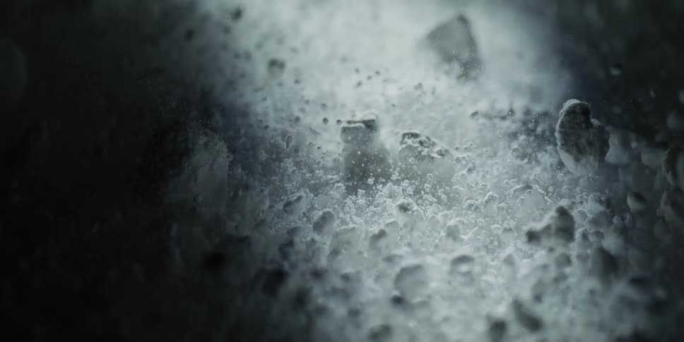 An abstract, slow-motion macro shot captures mysterious white particles flowing and tumbling in dramatic, moody lighting