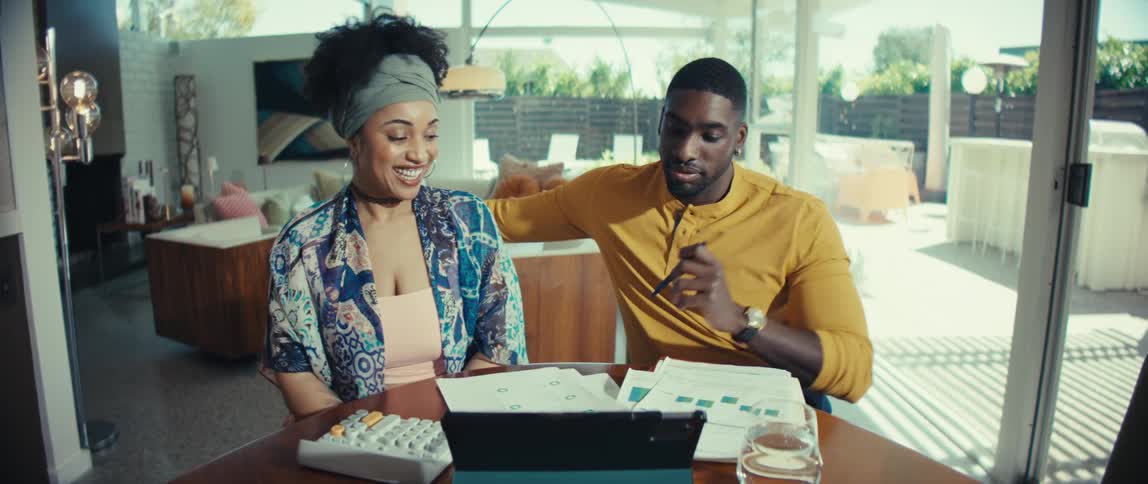 A young couple smiles while happily going over their finances and paperwork at a dining table in their sunny, modern home