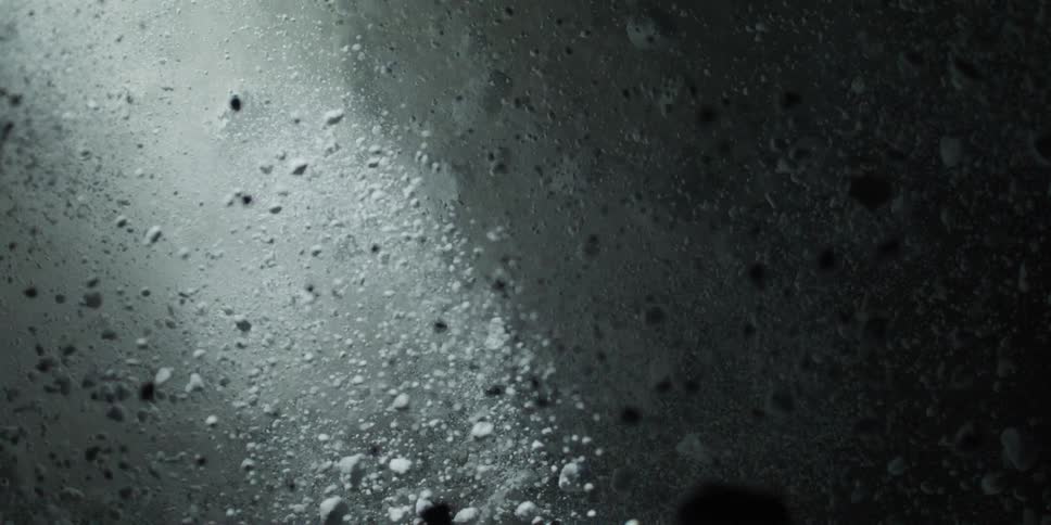 An abstract, high-contrast shot shows white, rocky particles floating and dispersing in slow motion