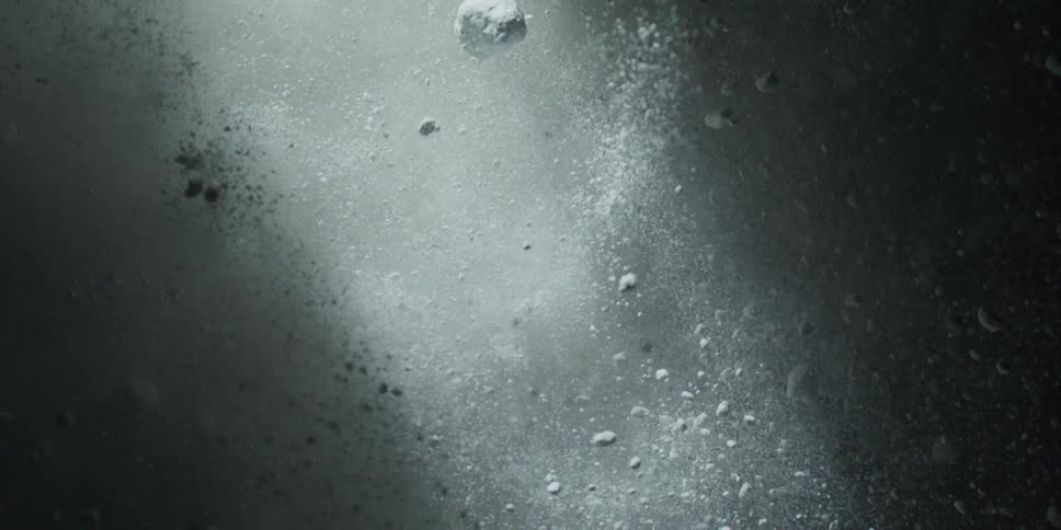 A dramatic slow-motion burst of white powder and particles expands against a dark background