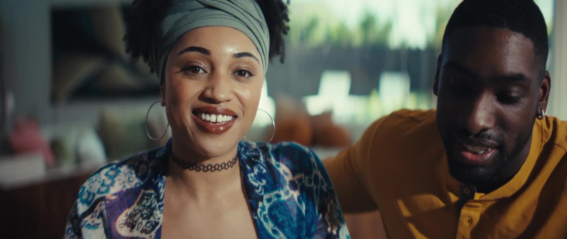 Close up of a beautiful young woman smiling and looking at the camera while sitting next to a man in a sunlit room