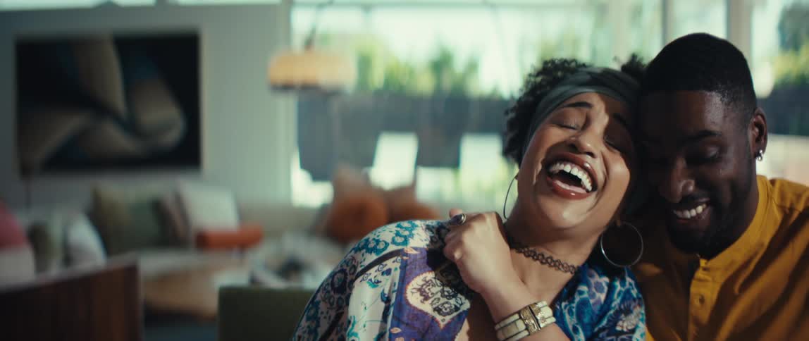 A stylish Black woman shares a joyful laugh with her partner while looking at something off-screen in their bright home