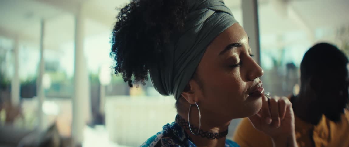 A beautiful, stylish woman wearing a headwrap sits thoughtfully in a sunlit room in a slow-motion close-up