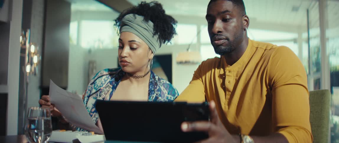 A young couple works together at home, seriously reviewing finances on a tablet and paper before smiling with success