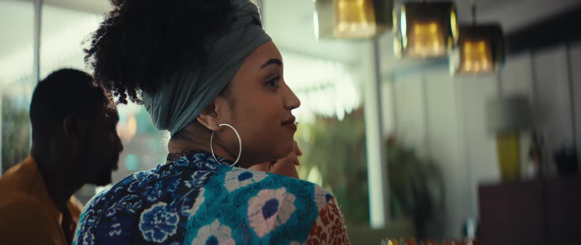 A stylish woman in a colorful top and headwrap has an animated conversation with a man before thoughtfully listening at a dining table