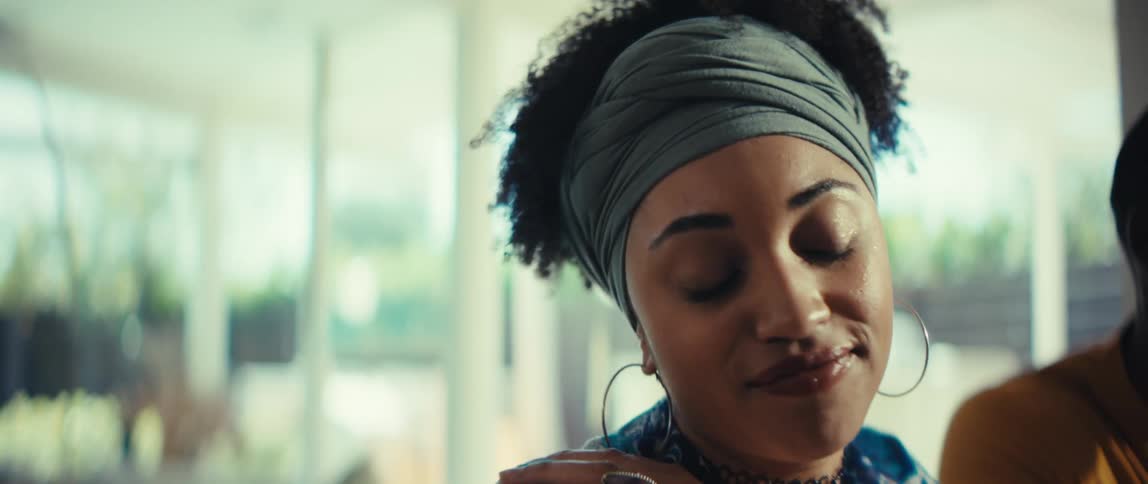 A stylish young woman with a headwrap sings and smiles while listening to music with her friend in a sunlit room