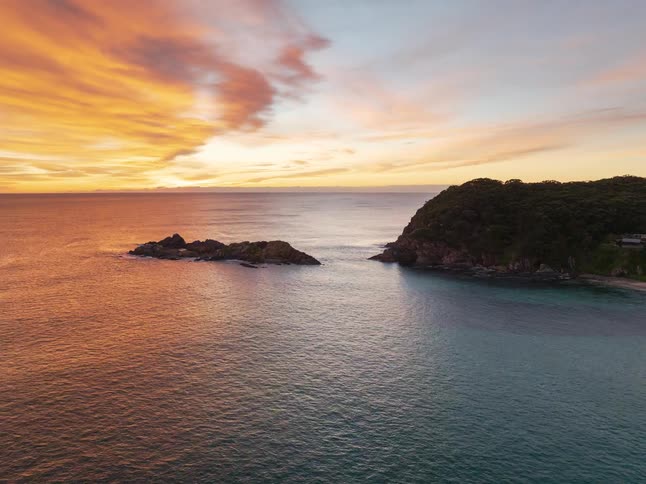 A breathtaking aerial shot pulls back from a fiery sunset reflecting on the ocean to reveal a tranquil island coastline