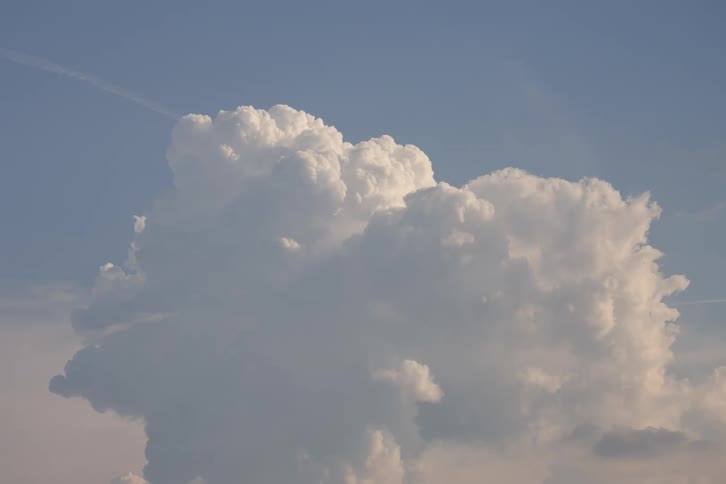 A large, puffy white cumulus cloud slowly grows and changes shape in a light blue sky as a jet flies by