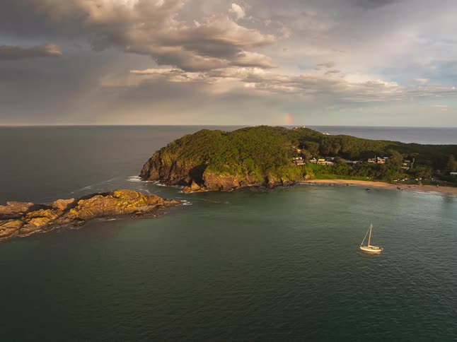 Aerial footage shows a sailboat anchored near a verdant headland with a lighthouse, while a rainbow glows under a stormy sunset sky