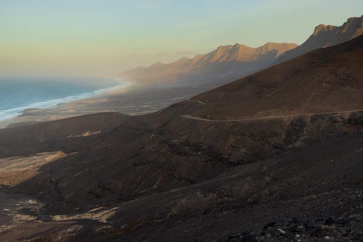A stunning day-to-night timelapse captures the dramatic landscape of a remote coastline, as cars drive along the winding mountain road during a vibrant sunset