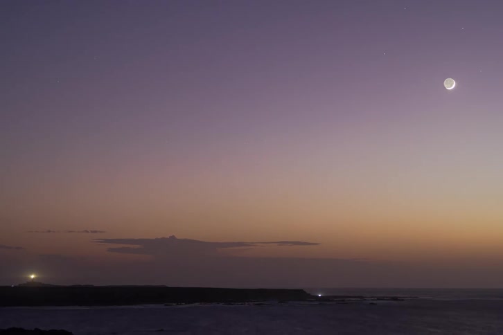 A timelapse from sunset to a starry night shows the moon's phases over a calm ocean with a flashing lighthouse on the horizon